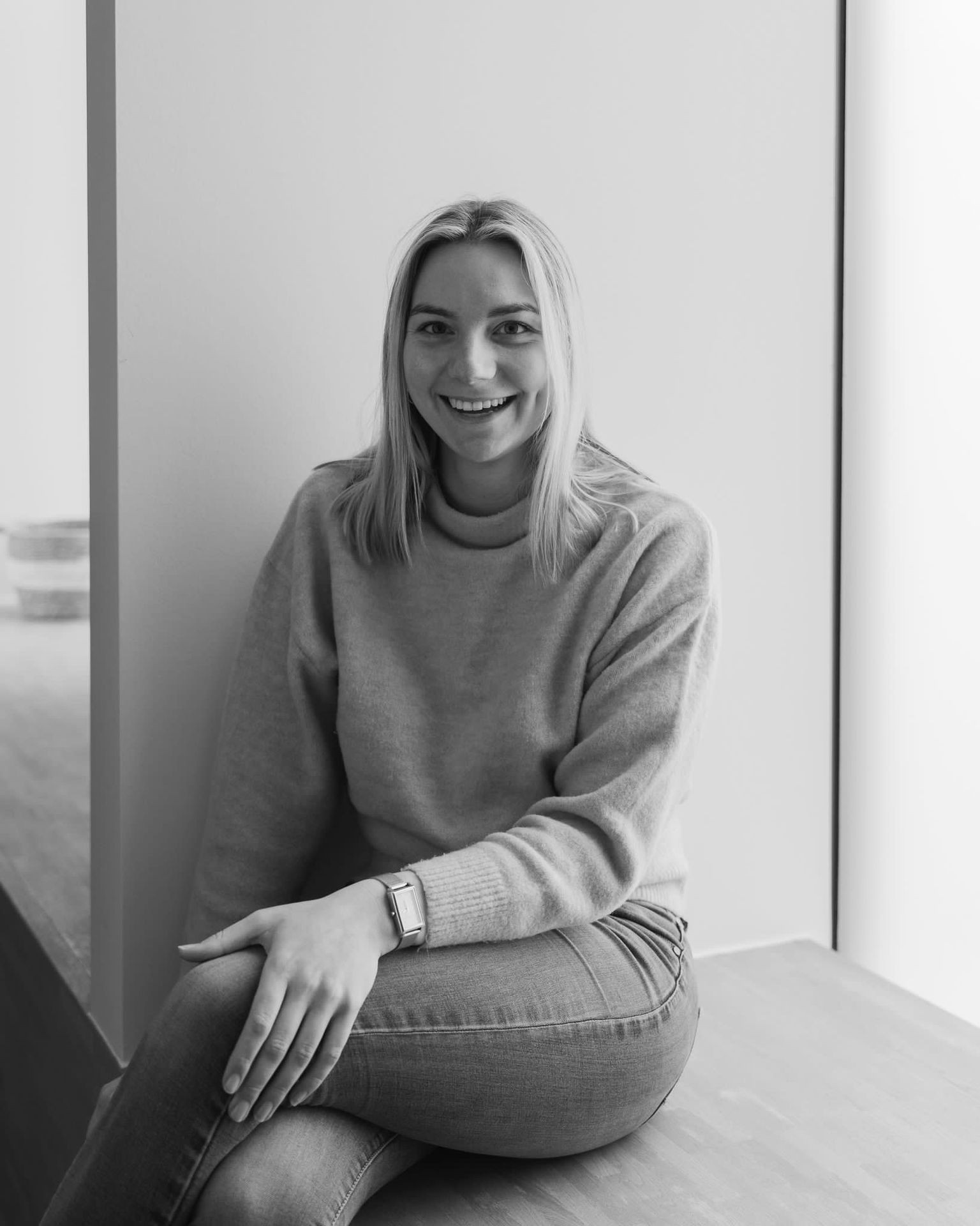 Person sitting by a wall, wearing a light sweater and jeans with a watch on their wrist.